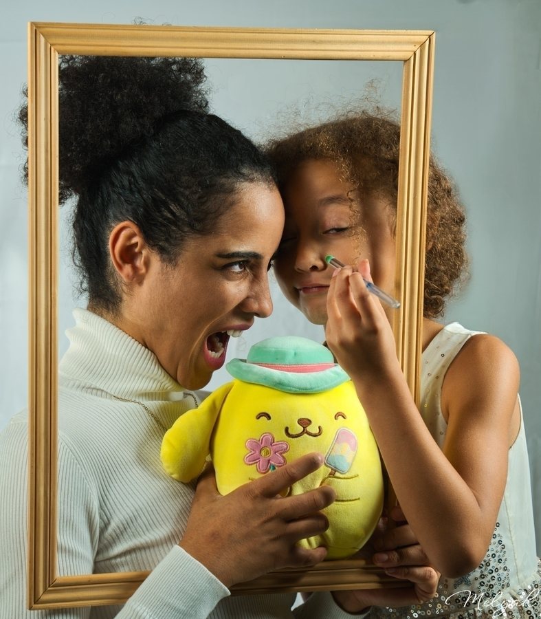 Une femme fait mine de croquer pendant qu'une fillette la maquille, une peluche jaune dans les bras